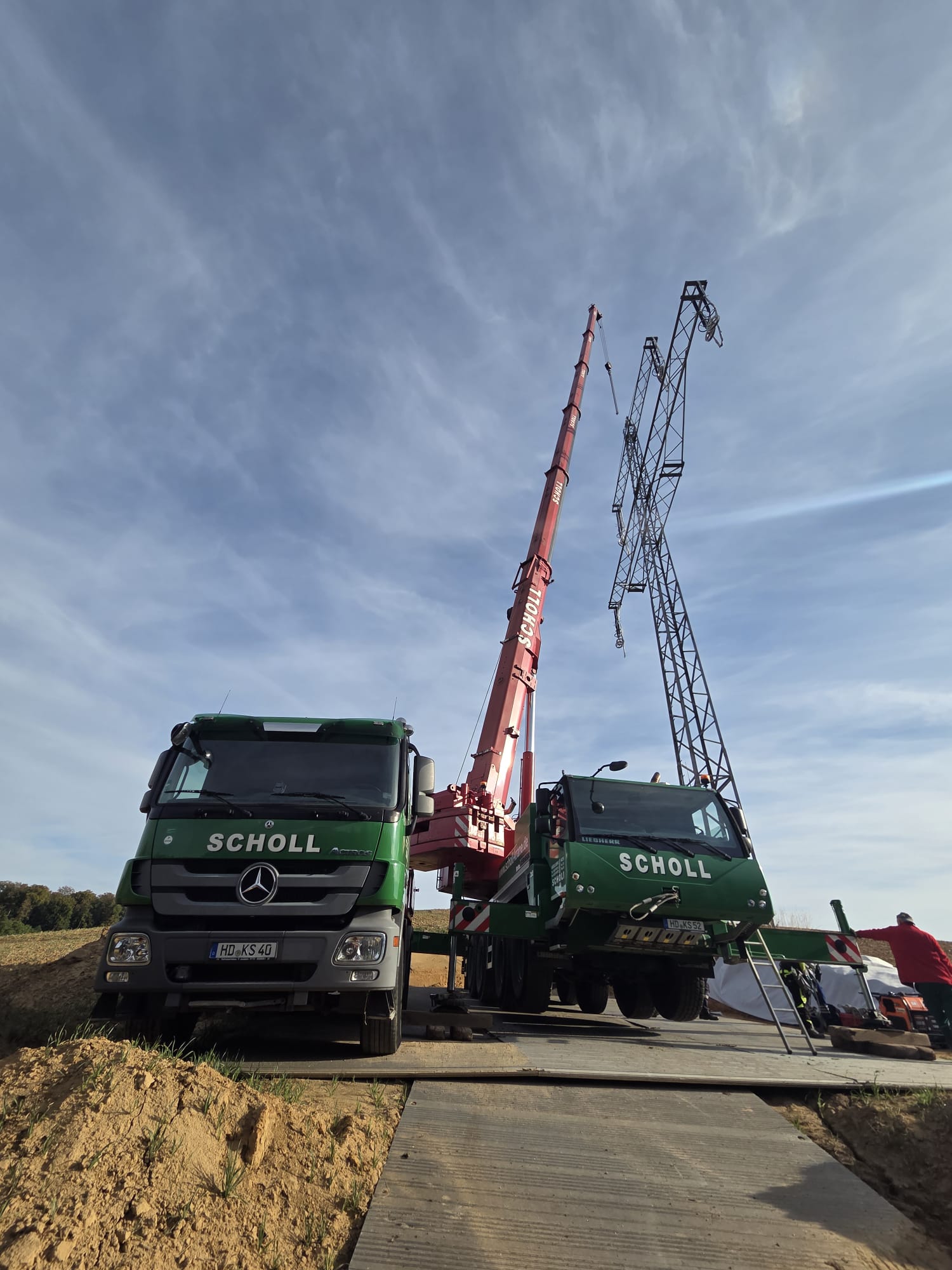 Autokran mit ausgefahrenem Ausleger auf einer Baustelle. Der Kran hebt eine Last an einem Seil vor blauem Himmel. Im Hintergrund sind weitere Baufahrzeuge, ein Gebäude und ein Bauzaun zu sehen.