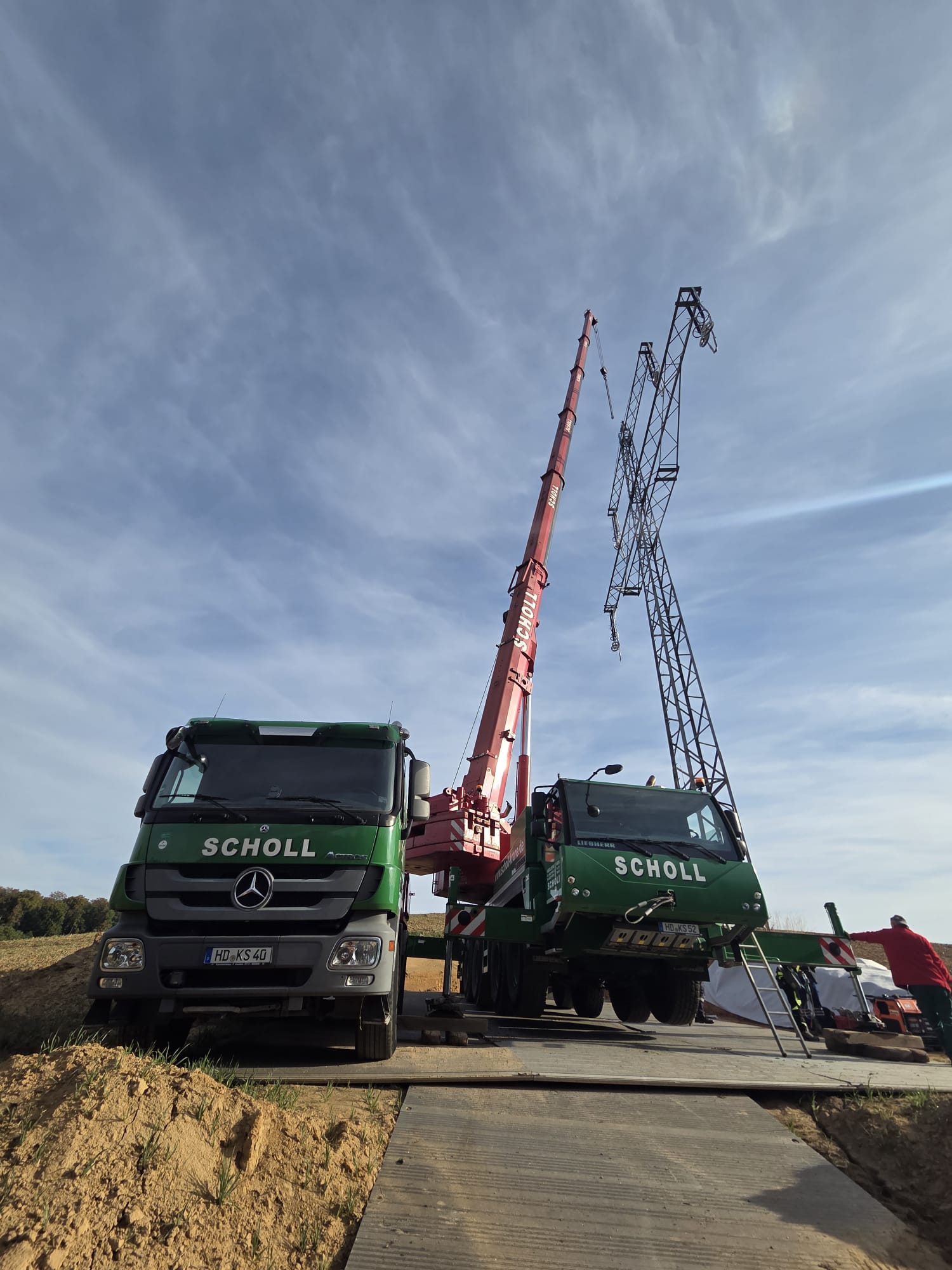 Autokran mit ausgefahrenem Ausleger auf einer Baustelle. Der Kran hebt eine Last an einem Seil vor blauem Himmel. Im Hintergrund sind weitere Baufahrzeuge, ein Gebäude und ein Bauzaun zu sehen.